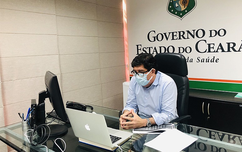 dr. cabeto sentado em frente computador usando máscara e com marca do governos do estado ao fundo